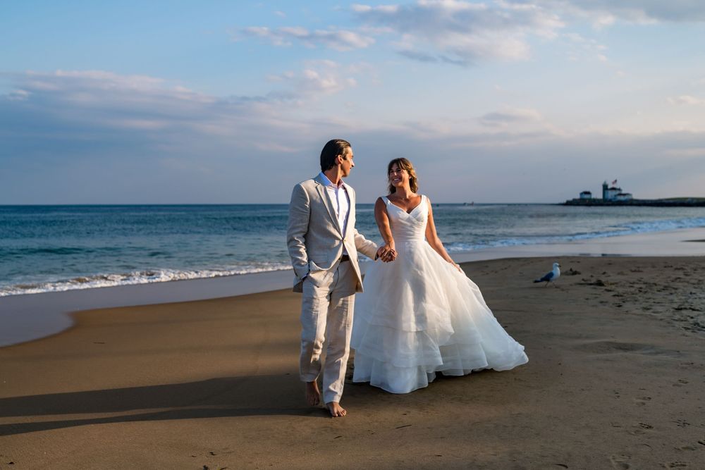 Bride and groom walk hand in hand along sandy beach at sunset.