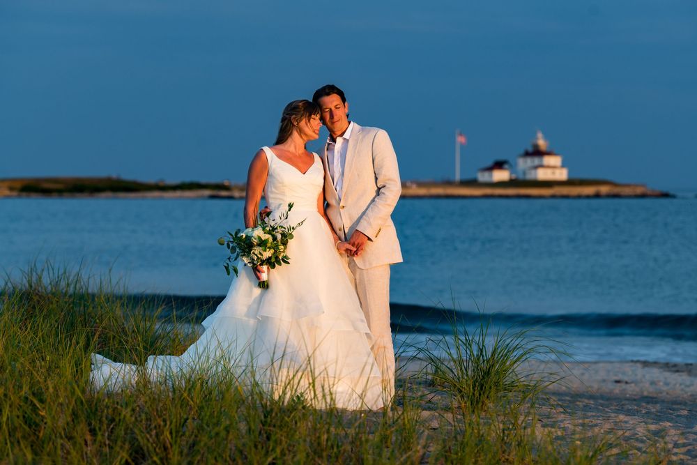 Couple embraces on beach with lighthouse in background at golden hour.