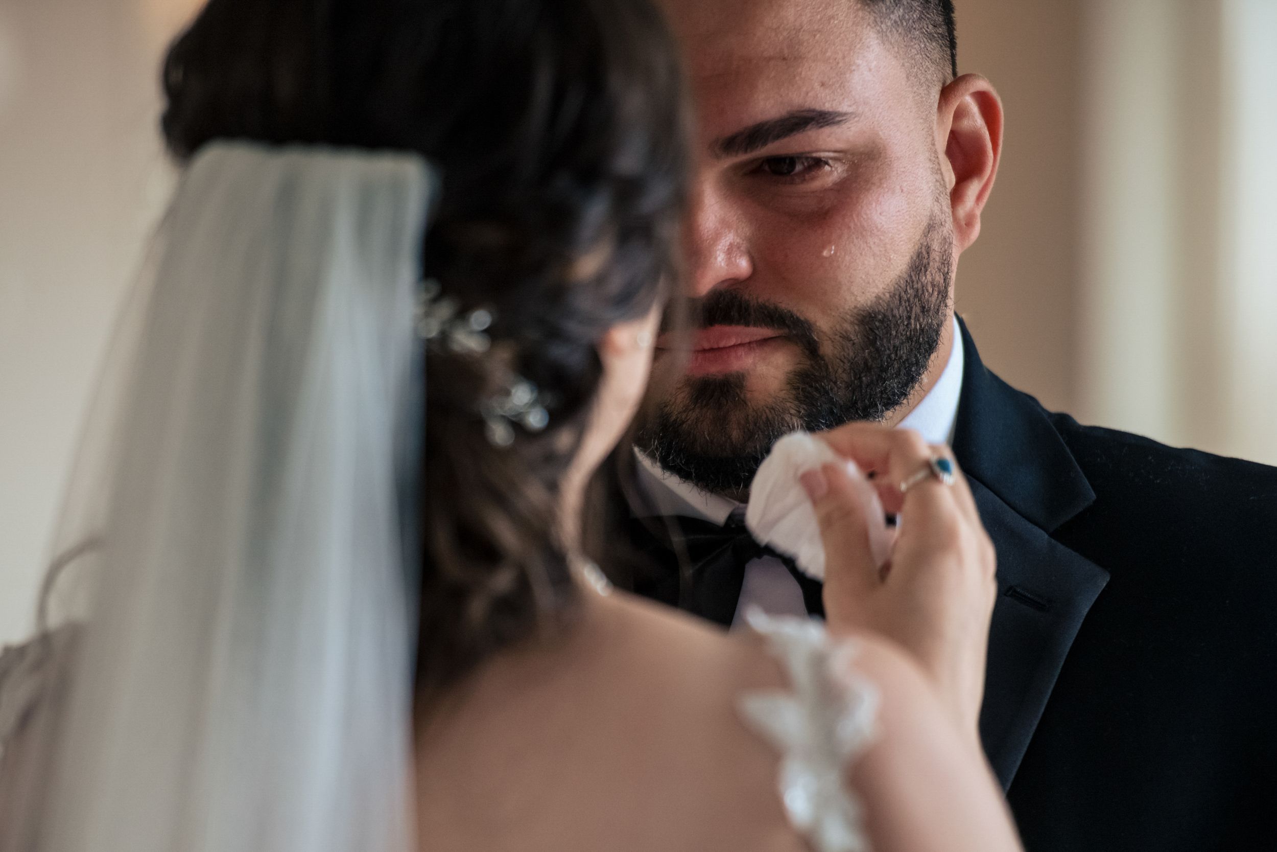 Emotional groom wiping tears during wedding ceremony as bride tenderly holds his face.