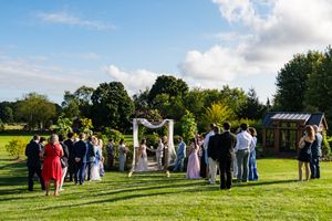 Outdoor wedding ceremony on green lawn with floral arch and guests gathered under blue sky.