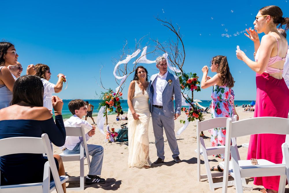 Beach wedding couple celebrating with guests blowing bubbles along sunny oceanside ceremony aisle.