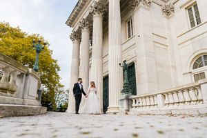 Elegant couple holding hands beside towering white columns of a historic Newport mansion.