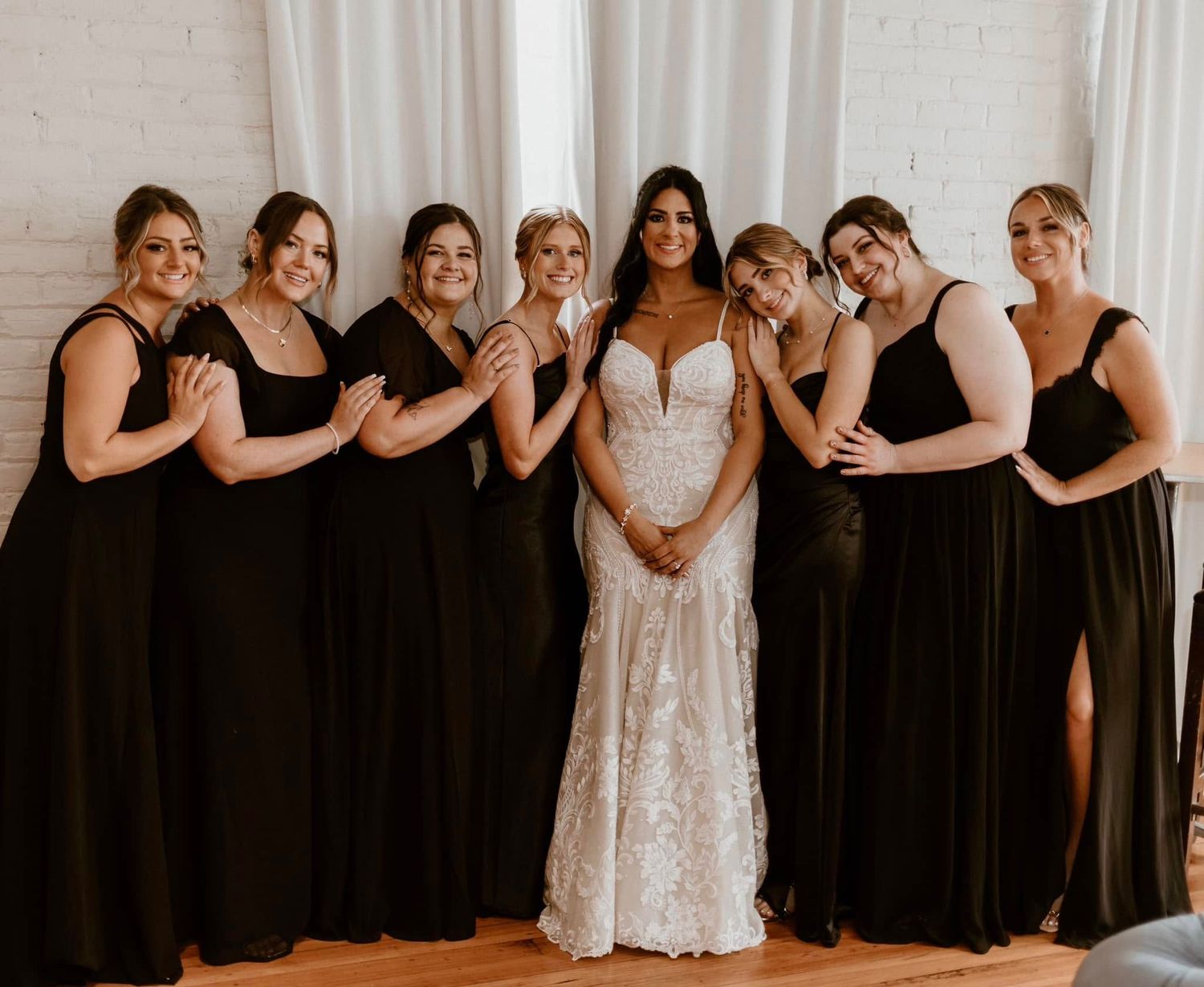 A wedding party in elegant black bridesmaid dresses poses with a bride in a white lace gown against a white backdrop.