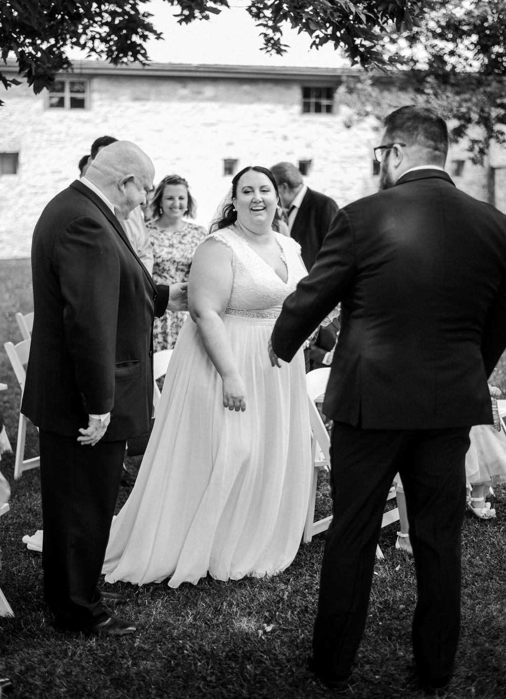Wedding party in formal attire arranges themselves on grass for group photos.