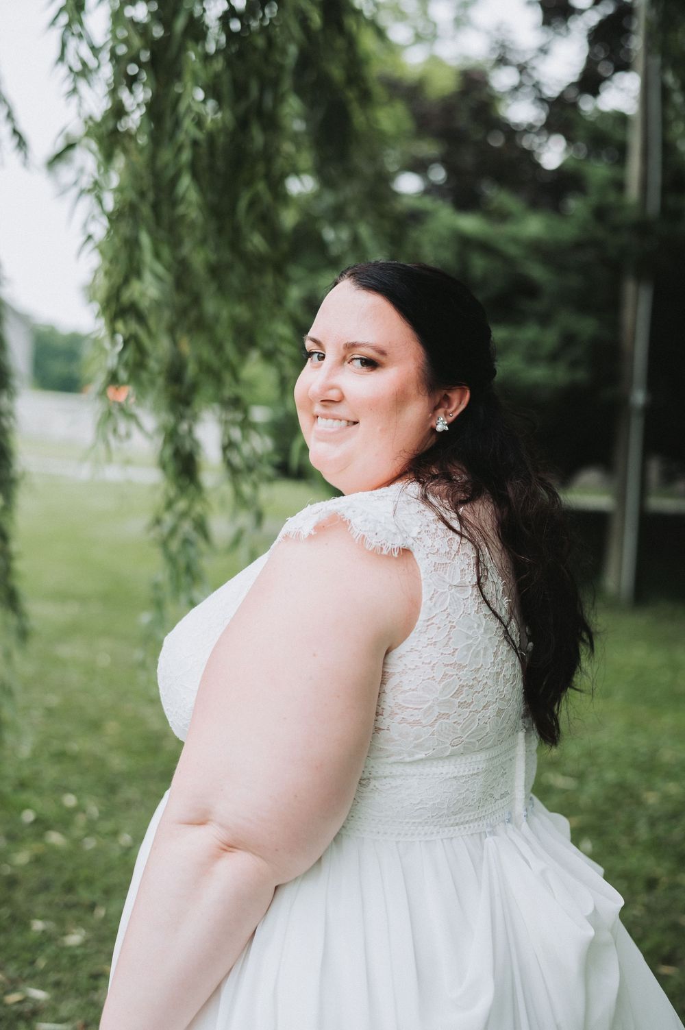 A bride in a lace wedding dress smiles while posing under willow trees.