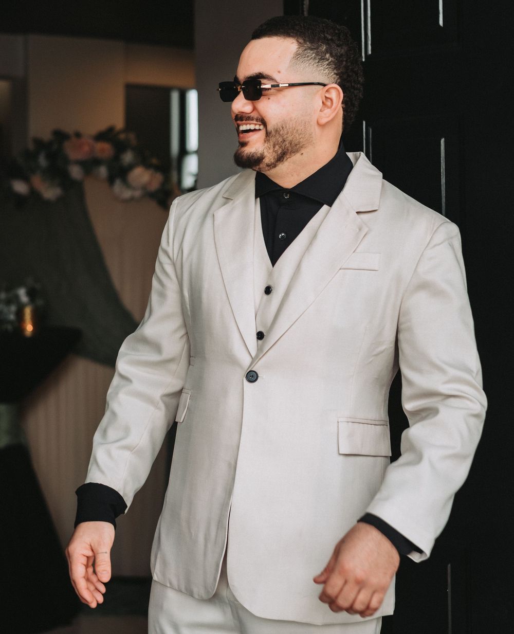 Smiling man in a light beige three-piece suit with black shirt and sunglasses at an elegant event.