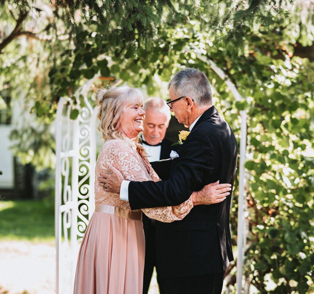 A couple shares an intimate moment during an outdoor wedding ceremony under green trees and white decor.