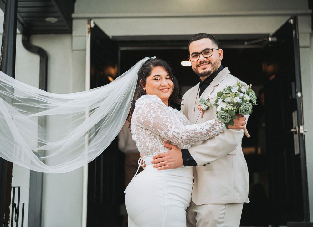 Bride in white lace gown with flowing veil embraces groom in beige suit holding bouquet at wedding venue entrance.