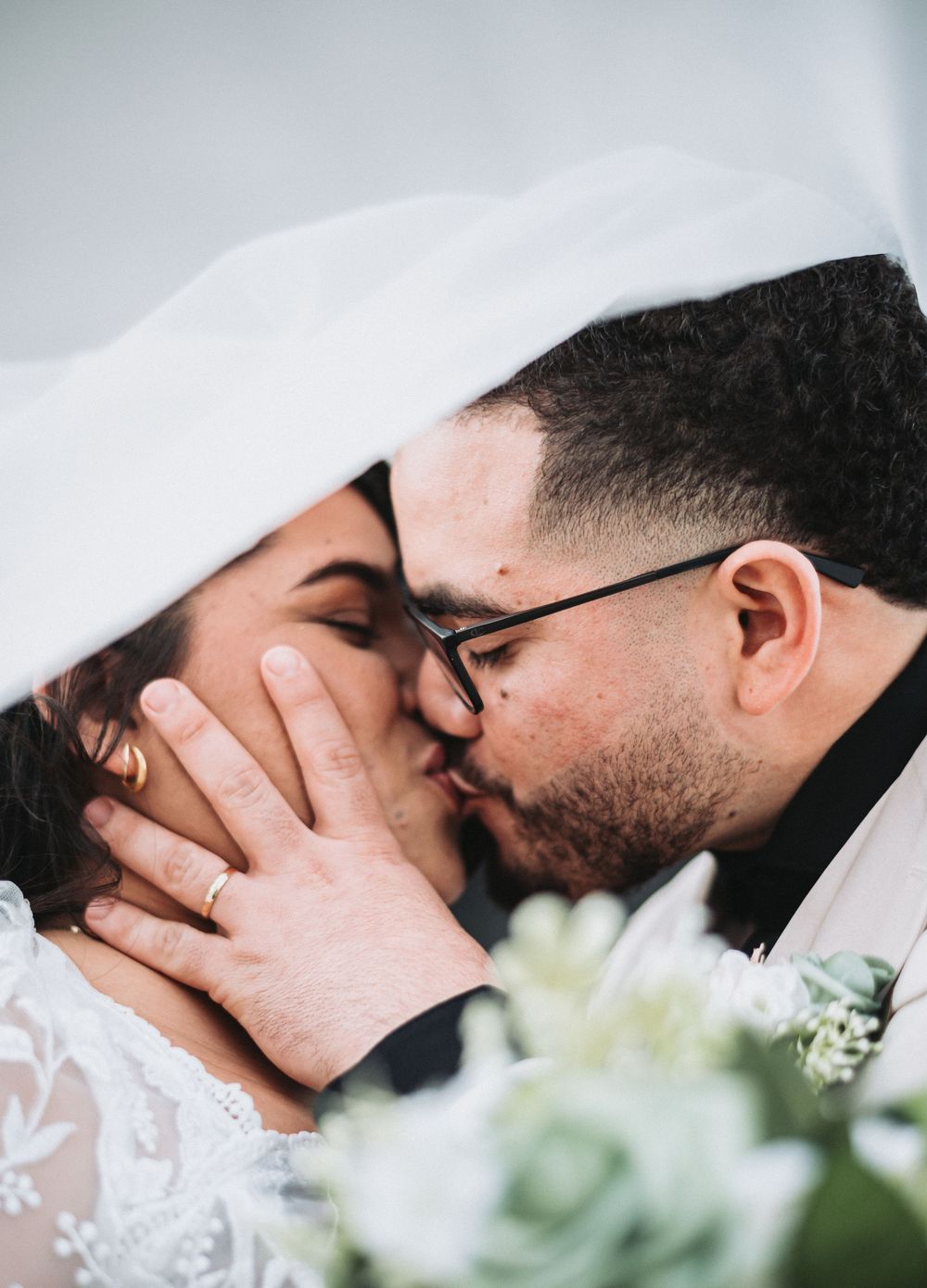A bride and groom share a romantic kiss under a white veil, with wedding flowers in the foreground.
