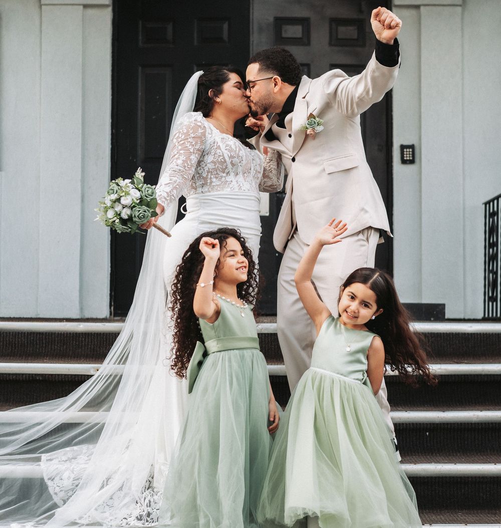 Bride and groom kissing on steps with two flower girls in sage green dresses celebrating at a wedding.