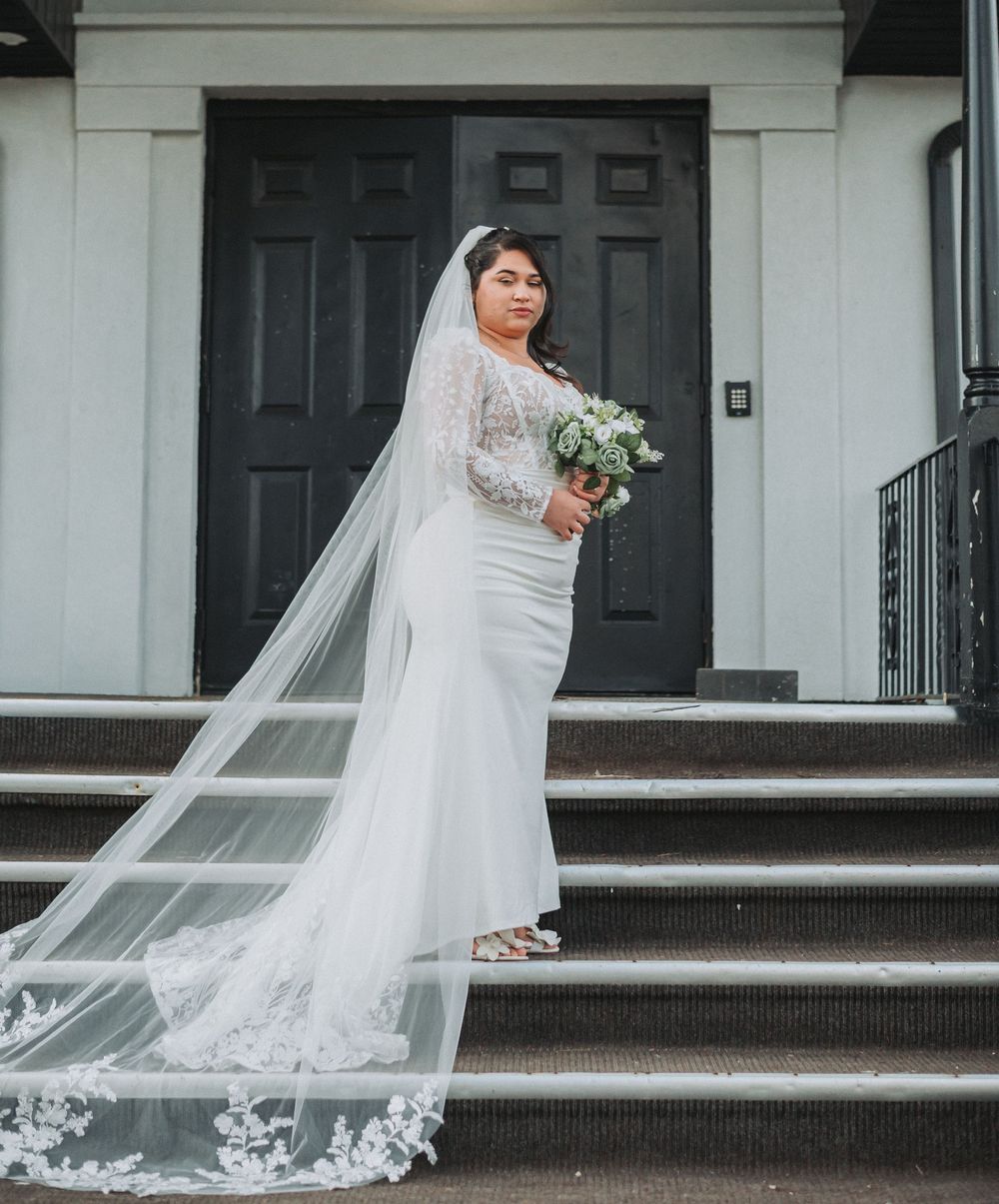 Bride in a white lace long-sleeve mermaid gown with cathedral veil holding bouquet on steps of elegant building.