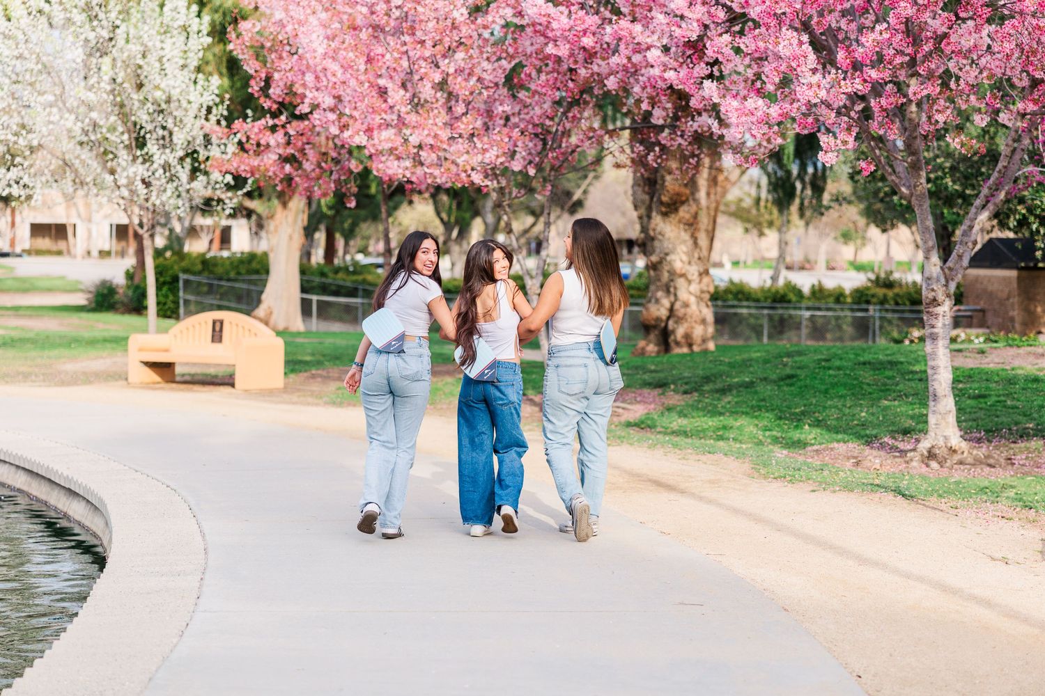 Three friends walk together under blooming pink cherry trees along a curved pathway in a spring park setting in Simi Valley CA