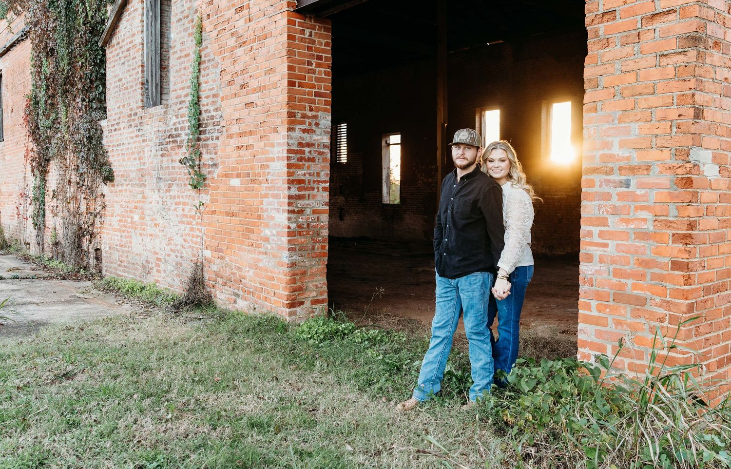 Two people stand in warm golden sunlight between brick archways of a rustic abandoned building with ivy growing on walls.