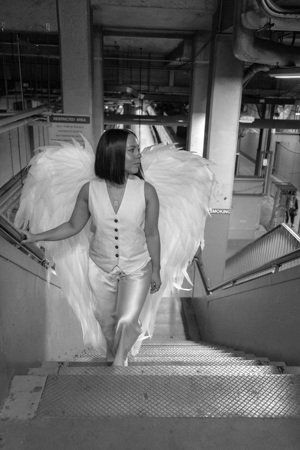 Figure in white with angel wings descending industrial metal stairs in black and white.