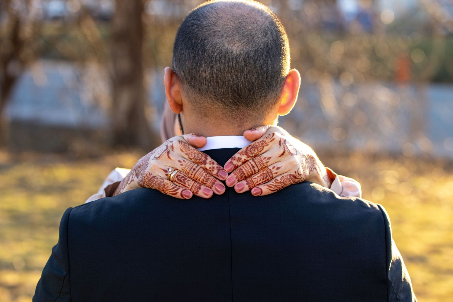 Hands with intricate henna designs rest on the back of someone wearing a black suit jacket outdoors.