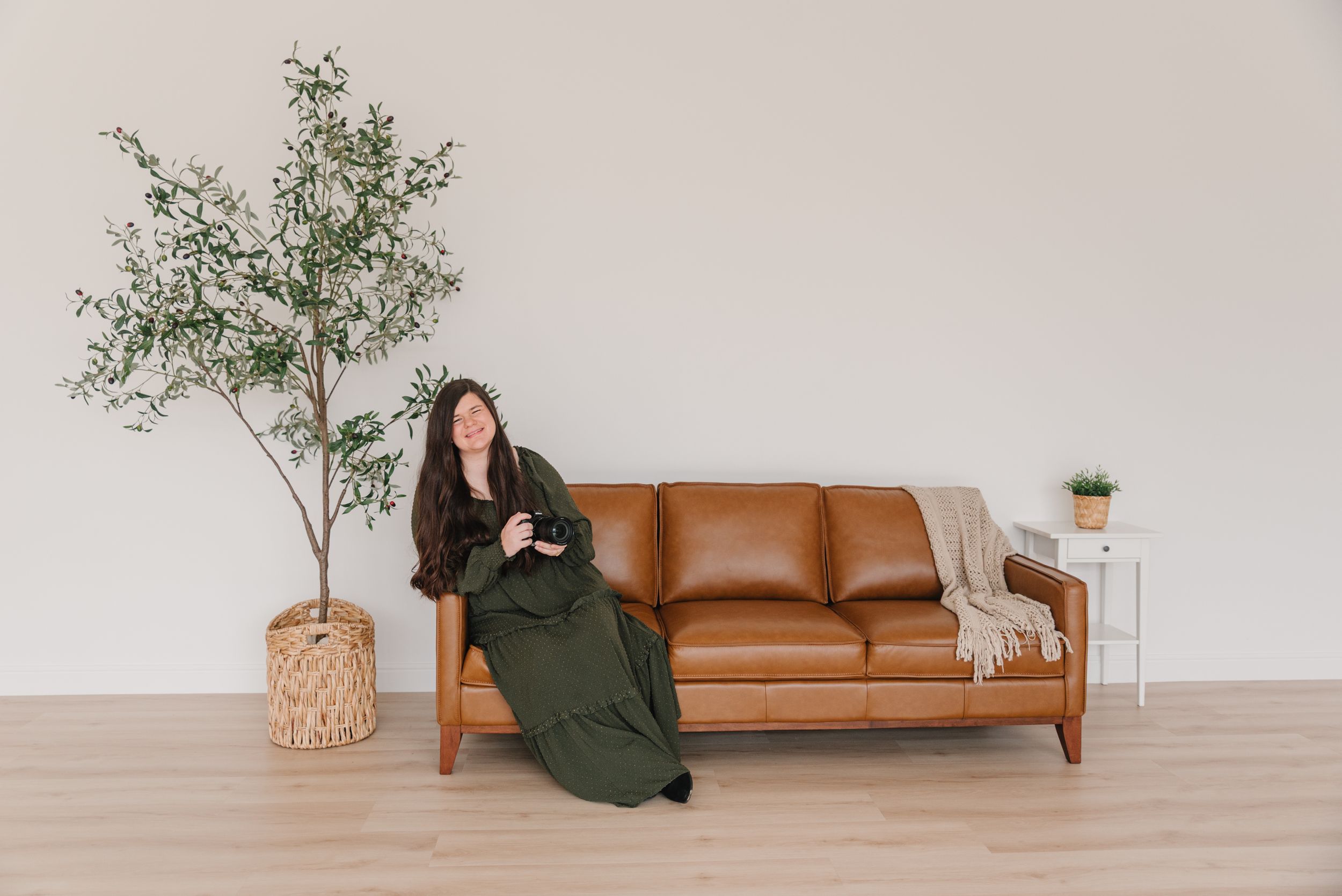 Contemporary living room features a tan leather sofa, potted olive tree, and someone relaxing in a green dress against white walls.