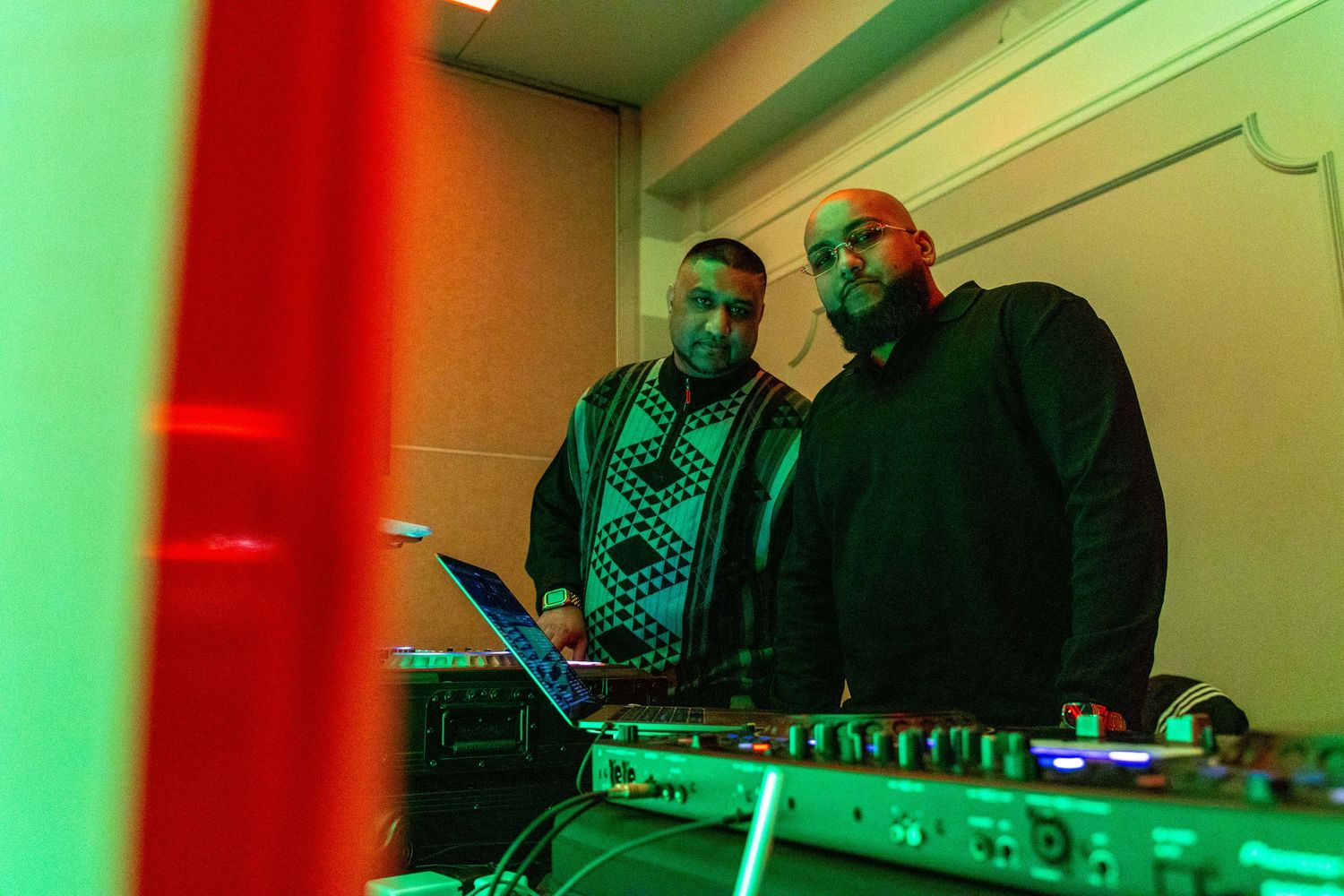 Two DJs perform behind mixing equipment under dramatic red and green lighting at a nightclub event.