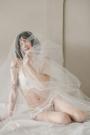 Artistic boudoir photo with white delicate tulle veil draping over figure in romantic lighting against neutral backdrop.
