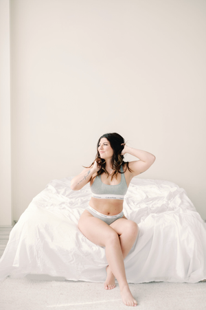 A figure in gray lingerie sits gracefully on white bedding in a minimalist setting with natural lighting.