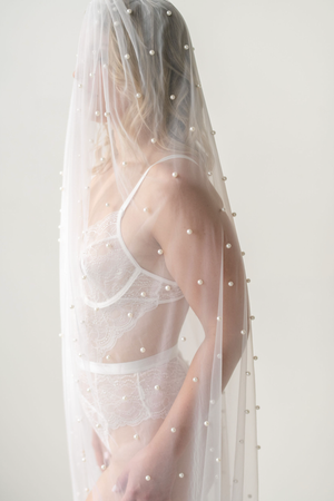 Ethereal silhouette behind sheer white fabric decorated with pearl embellishments and soft lighting.