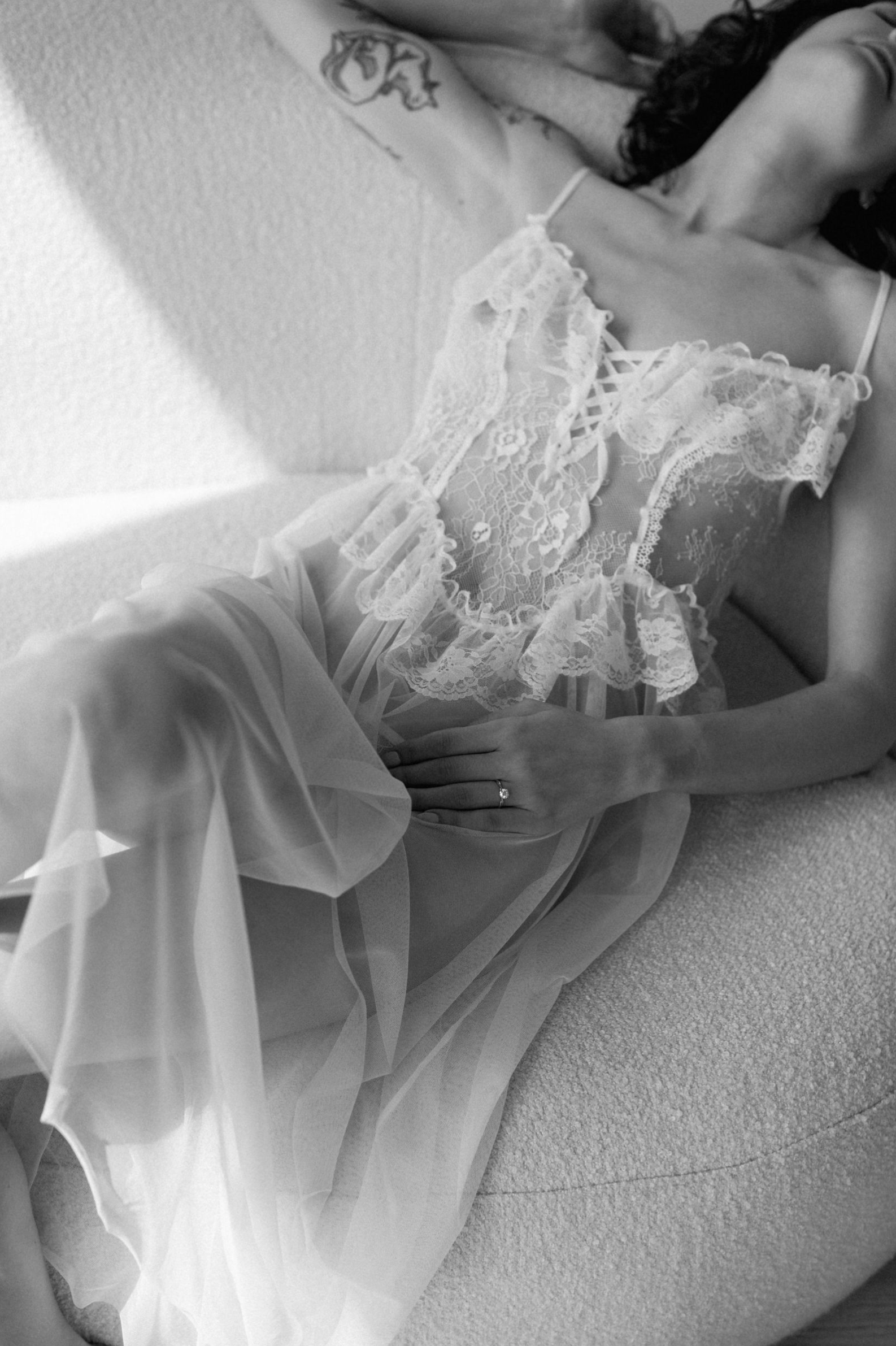 A black and white boudoir photograph featuring delicate white lace lingerie and flowing sheer fabric in soft lighting.