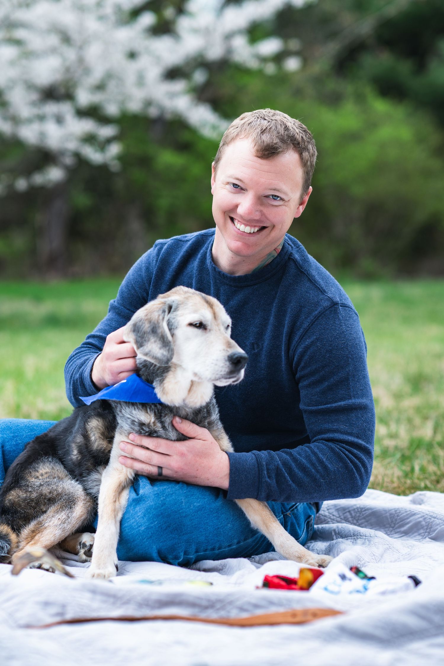 A person in a blue sweater sits on a blanket outdoors while petting a senior husky-mix dog with gray fur.