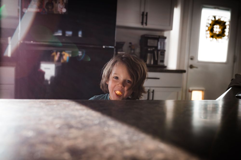 A child peeks over a dark countertop in a sunlit kitchen with a decorative wreath visible in the background.