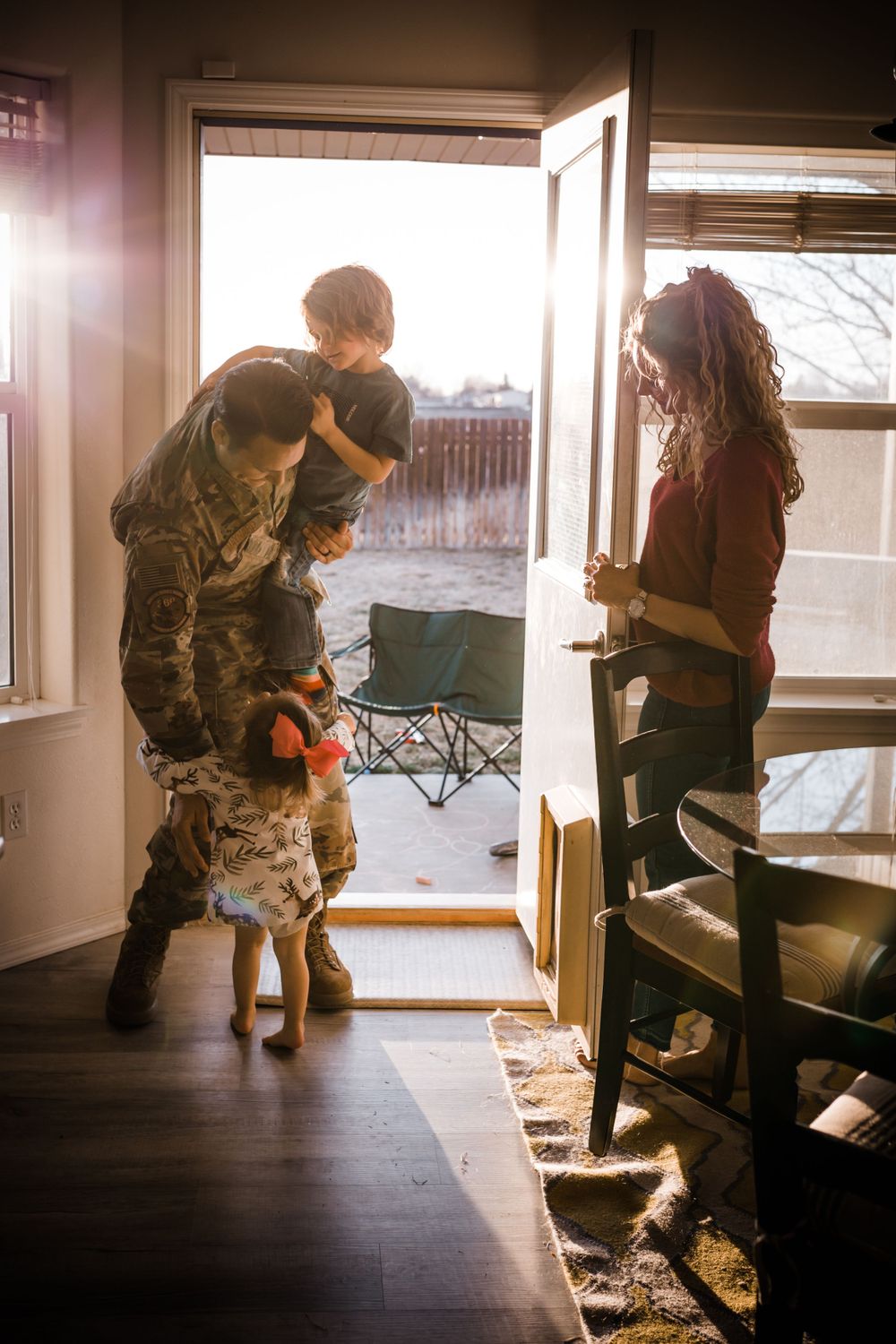 A touching indoor scene in warm lighting shows a military service member in camouflage greeting a dog near an open doorway.