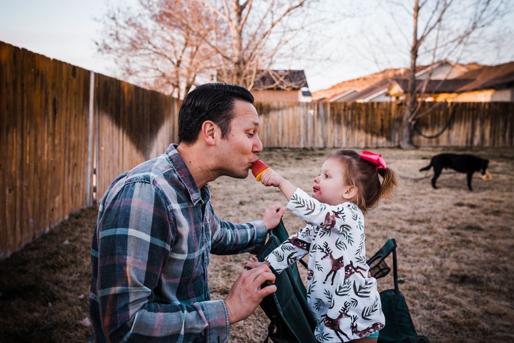 An adult and young child share a playful moment in a backyard with wooden fencing at sunset while a dog walks by.