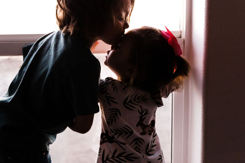 Silhouetted figures of siblings sharing a sweet moment in front of a sunlit window.