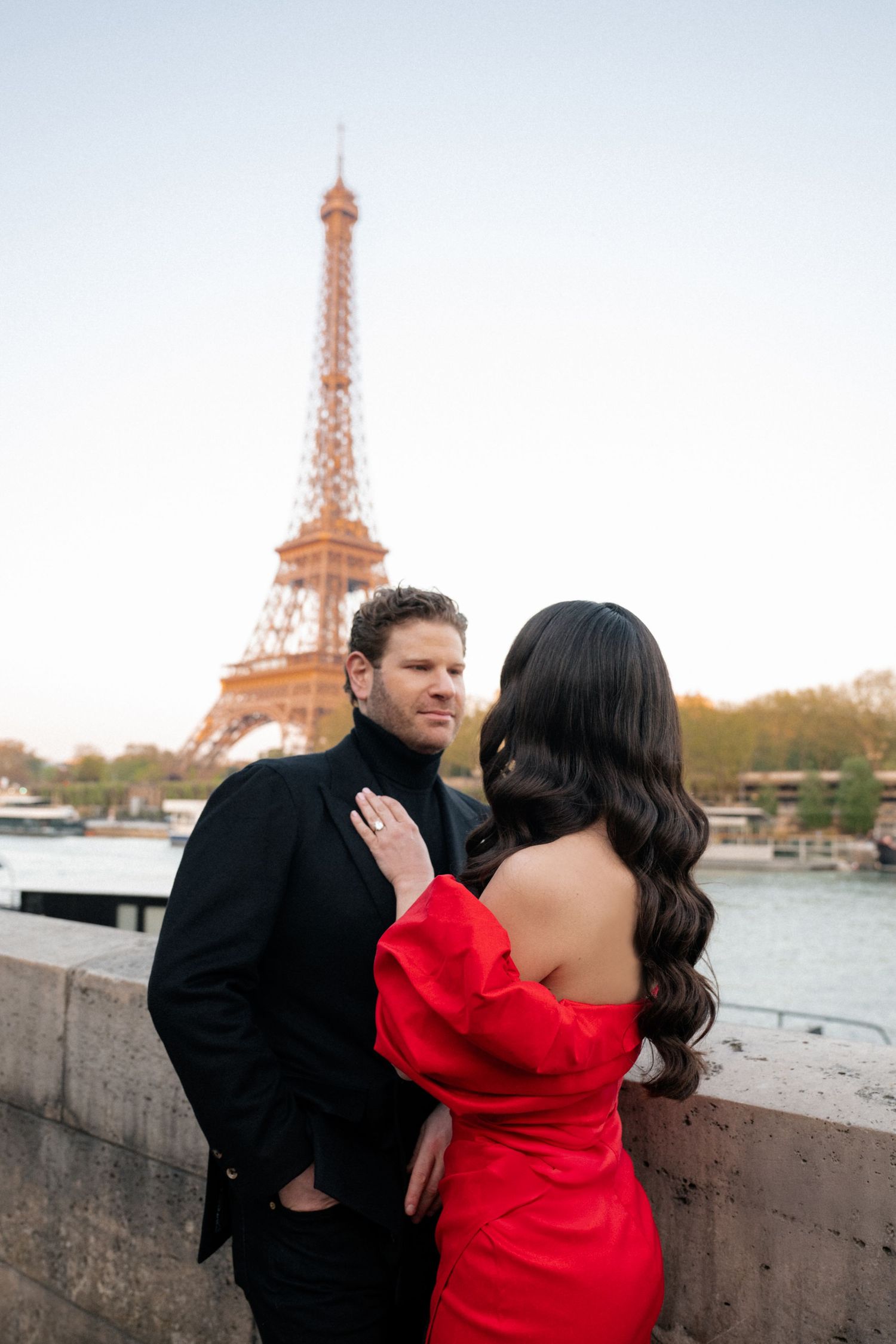A couple stands together along the Seine river with the Eiffel Tower in the background at sunset.