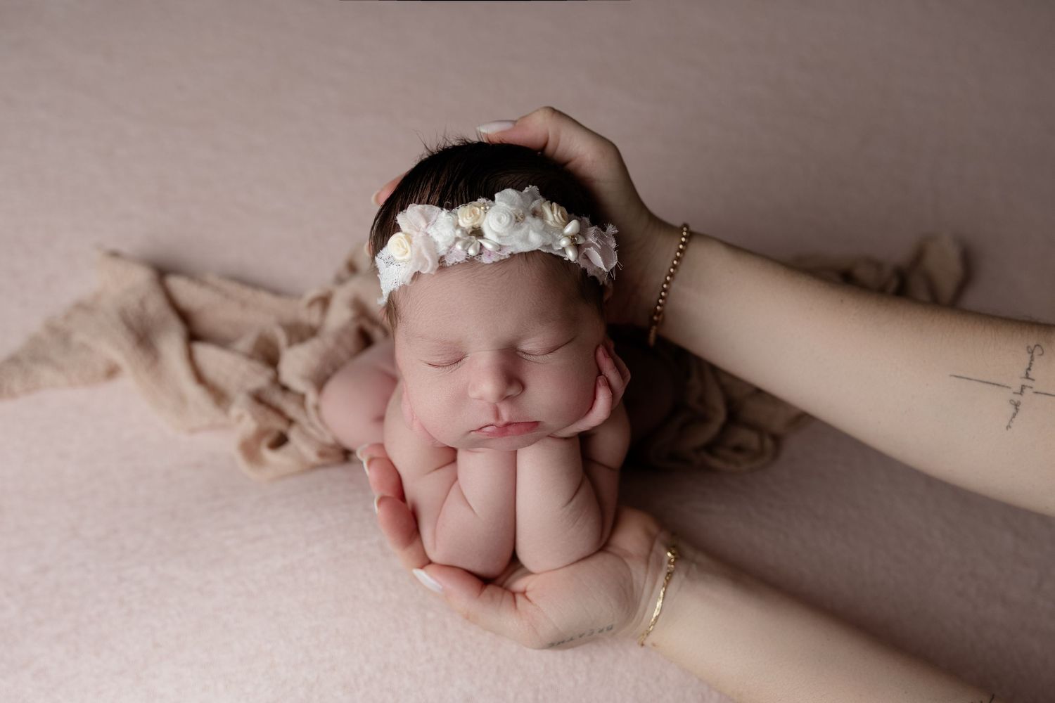 Sleeping newborn wearing a delicate floral headband rests peacefully on hands against pink fabric background.
