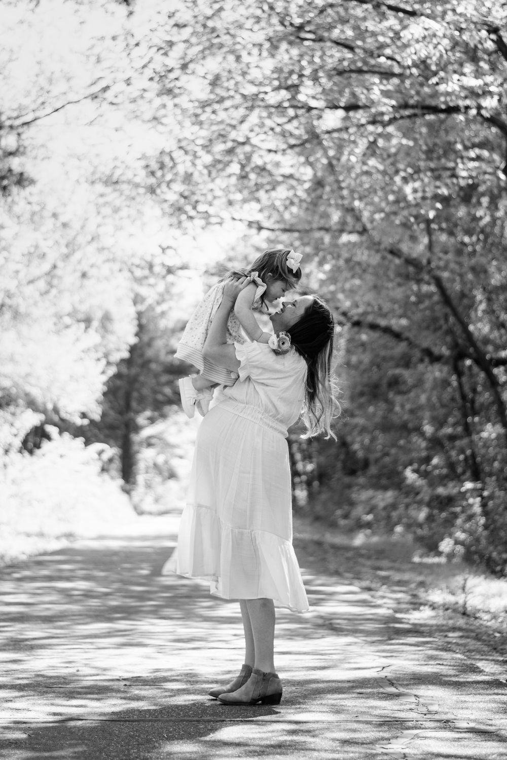 A tender moment captured in black and white as a figure lifts a small child on a tree-lined path.