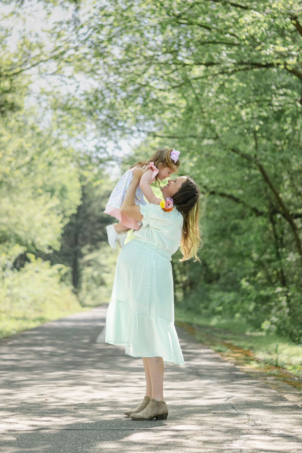 A parent in a mint green dress lifts a small child on a tree-lined path in a sunlit outdoor setting.