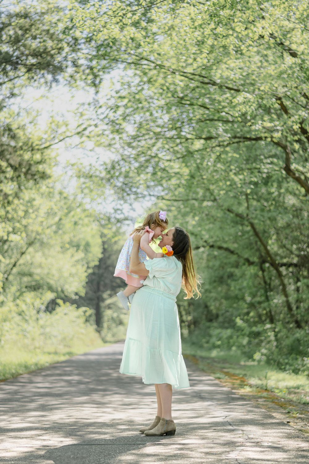 A figure in a mint green dress lifts a small child along a tree-lined pathway on a bright sunny day.