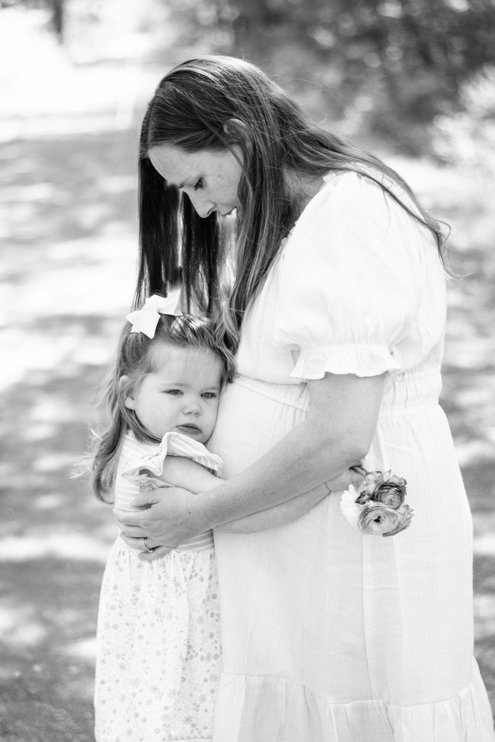 A tender black and white portrait of a parent and child embracing outdoors while wearing white dresses.