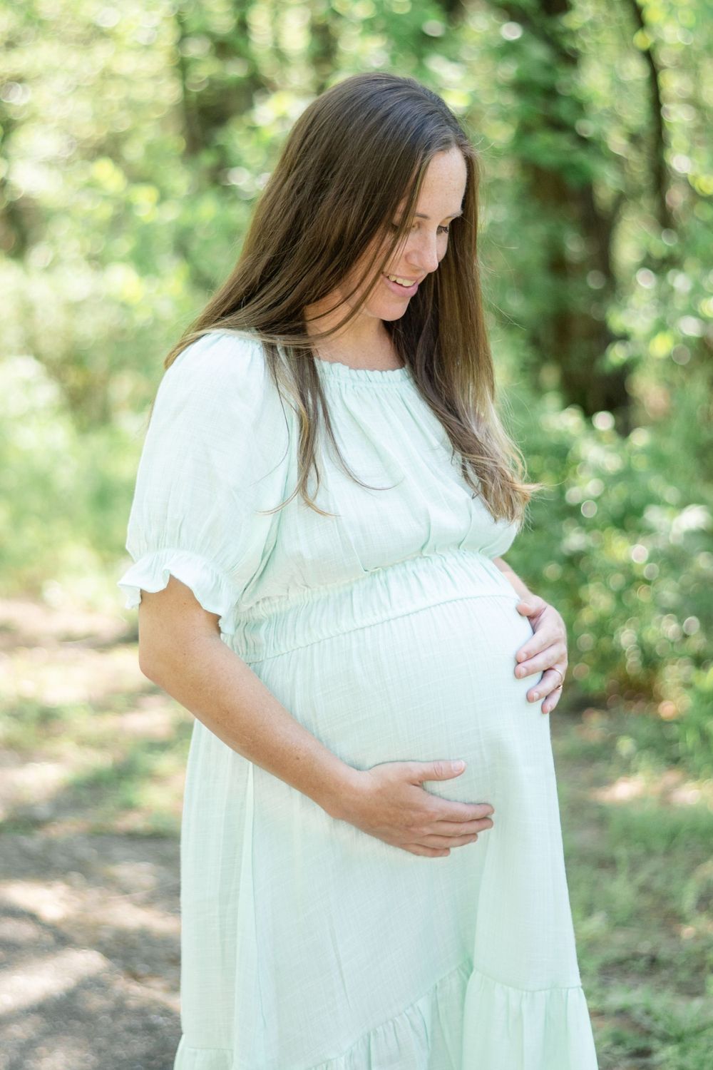 A woman in a flowing white dress cradles her pregnant belly while standing on a sunlit forest path.