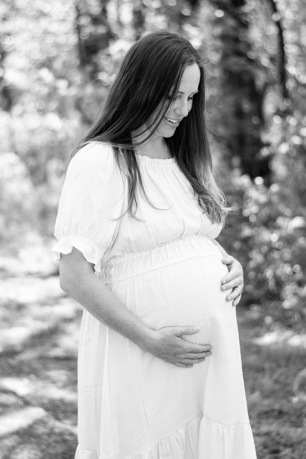 Black and white photo of a pregnant person in a white dress standing outdoors in a natural setting.
