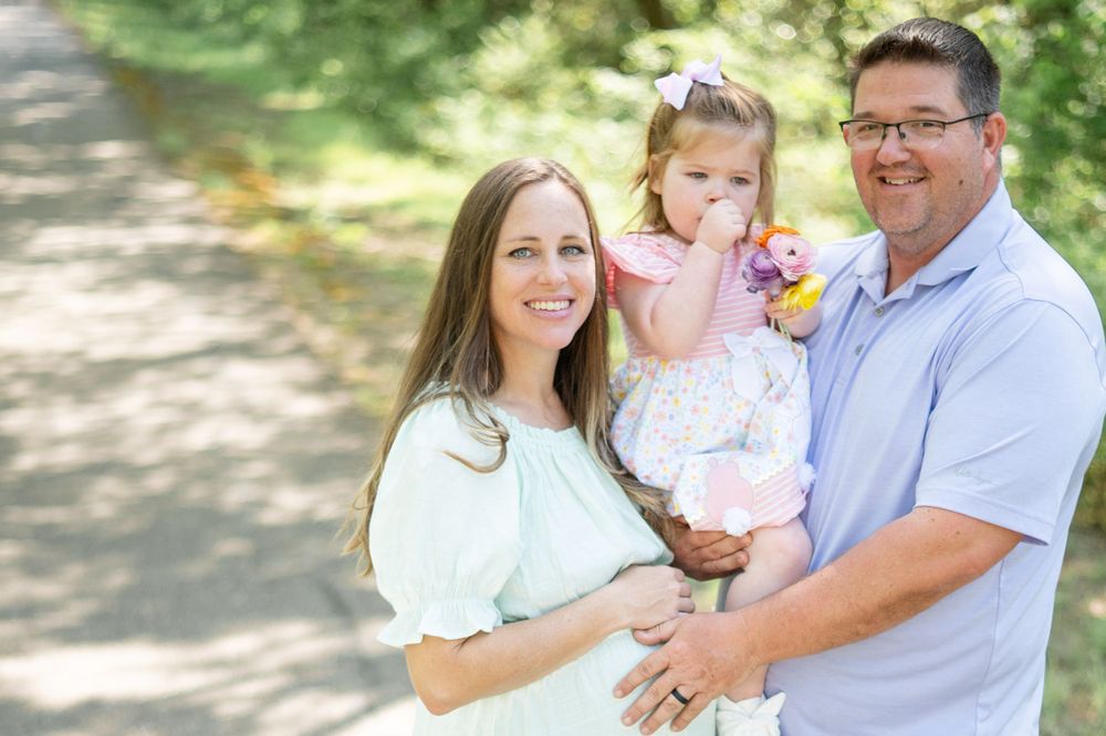 A family smiling together outdoors on a sunny day along a tree-lined path.