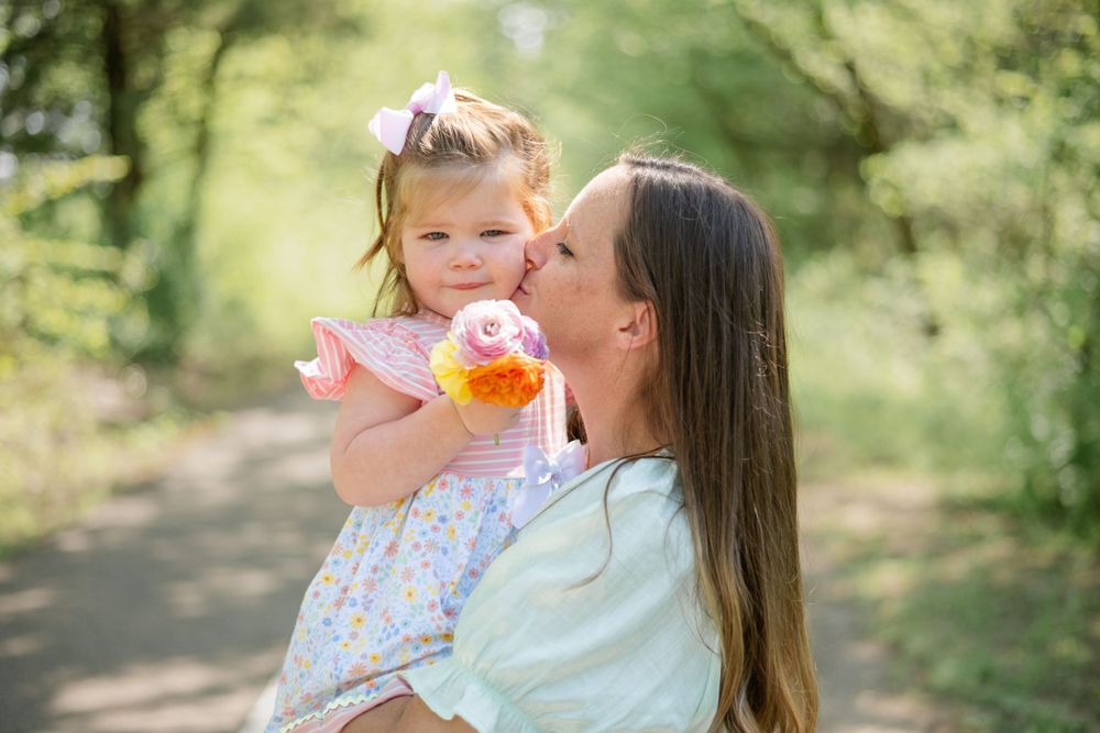 A loving moment shared between parent and child on a sunlit forest path, with a pink floral dress and soft smiles.