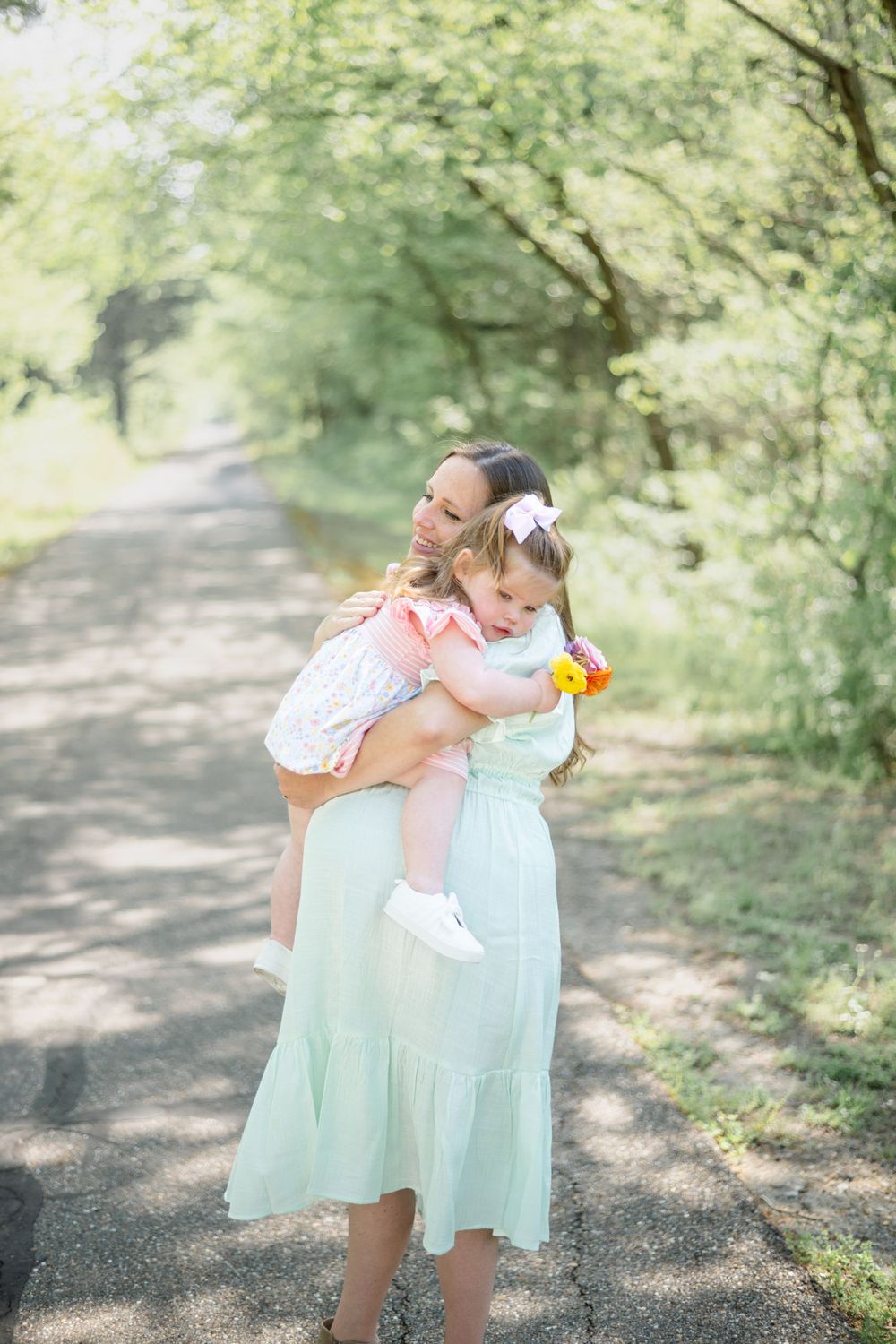 A gentle nature path surrounded by green trees creates a dreamy backdrop for two figures in soft pastel dresses.