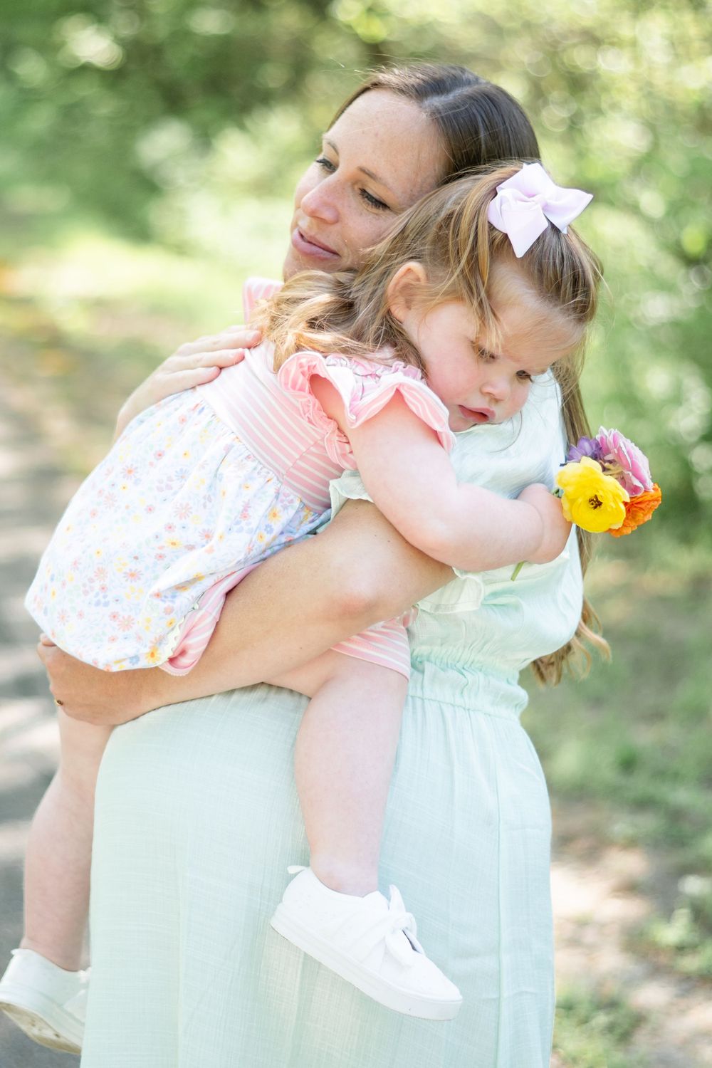 A tender moment captured outdoors as a woman in a mint dress holds a small child wearing pink and white with a hair bow.