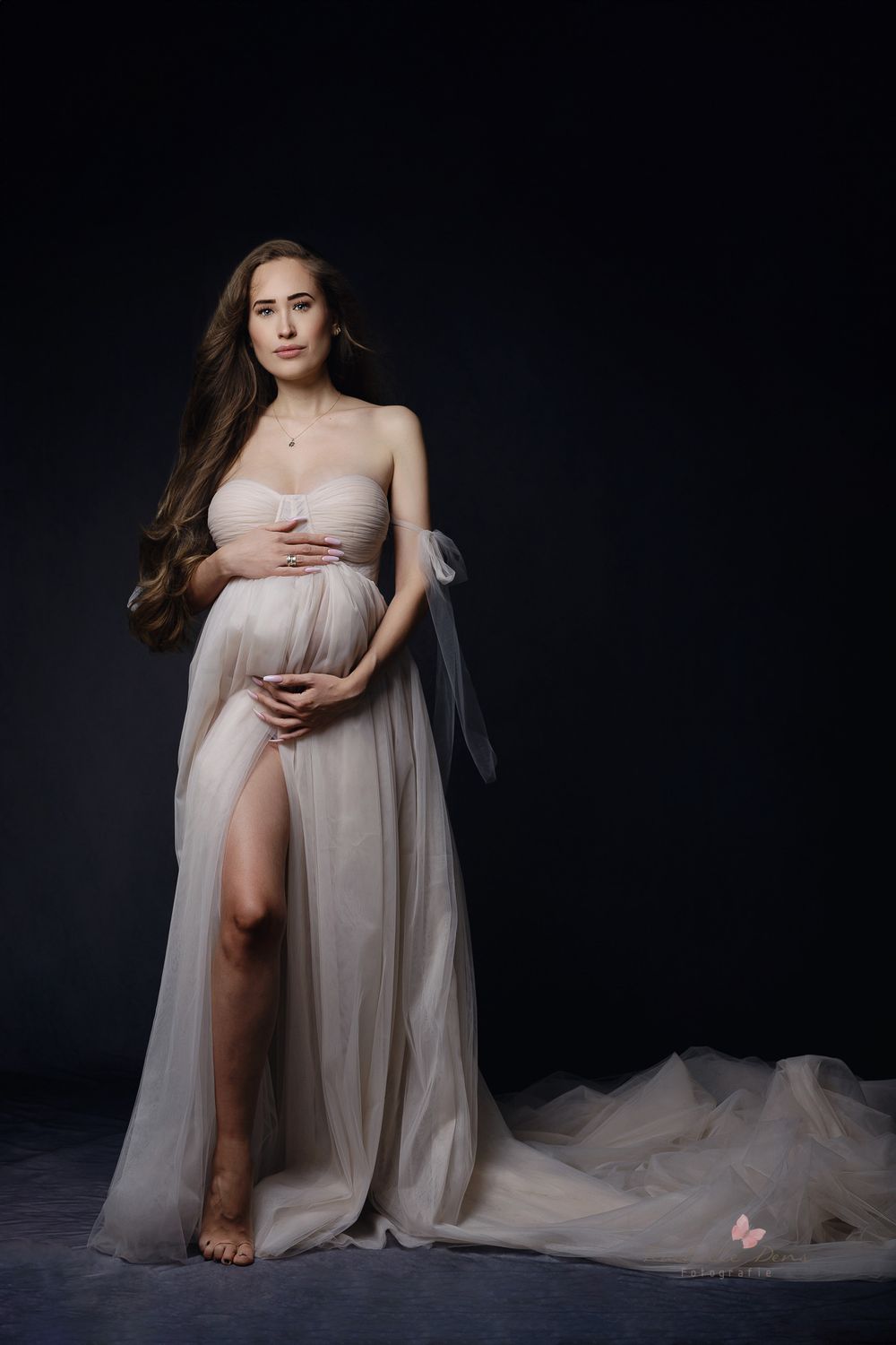 A dramatic maternity portrait in a flowing blush gown against a dark backdrop with ethereal lighting and long train.
