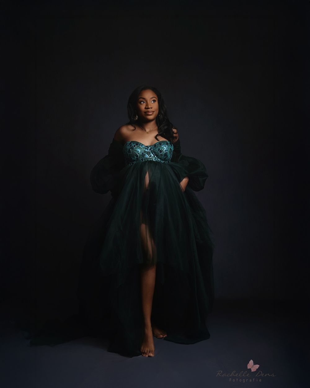 Glamorous portrait against dark backdrop features elegant emerald green flowing gown with dramatic tulle and sequin details.