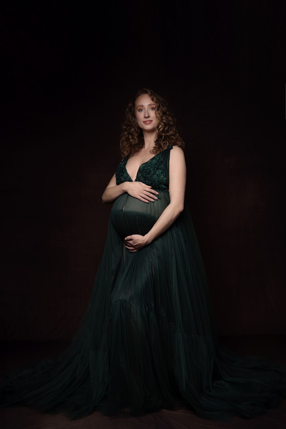 A maternity portrait featuring an elegant emerald green flowing gown against a dark background in studio lighting.