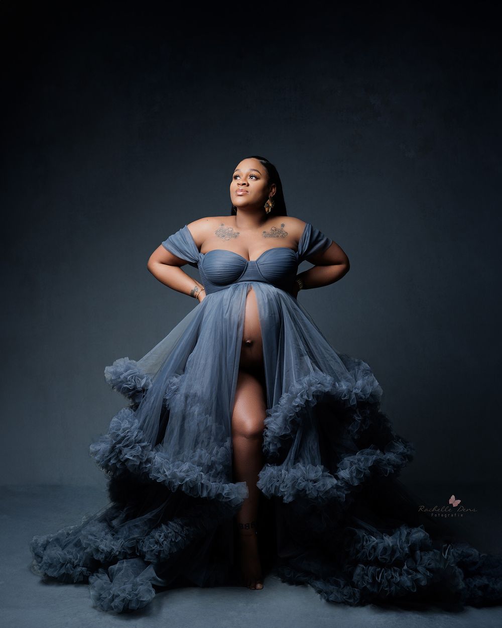 Elegant maternity portrait of a person in a flowing dusty blue tulle gown with dramatic ruffles against dark backdrop.