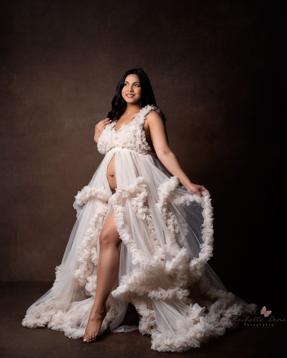 A flowing white tulle gown with floral appliques photographed against a dark brown studio backdrop.