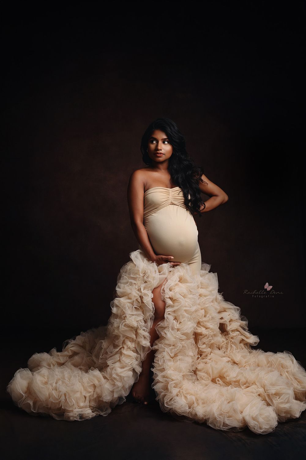 A stunning maternity photo featuring a dramatic ruffled champagne-colored gown against a dark studio backdrop.