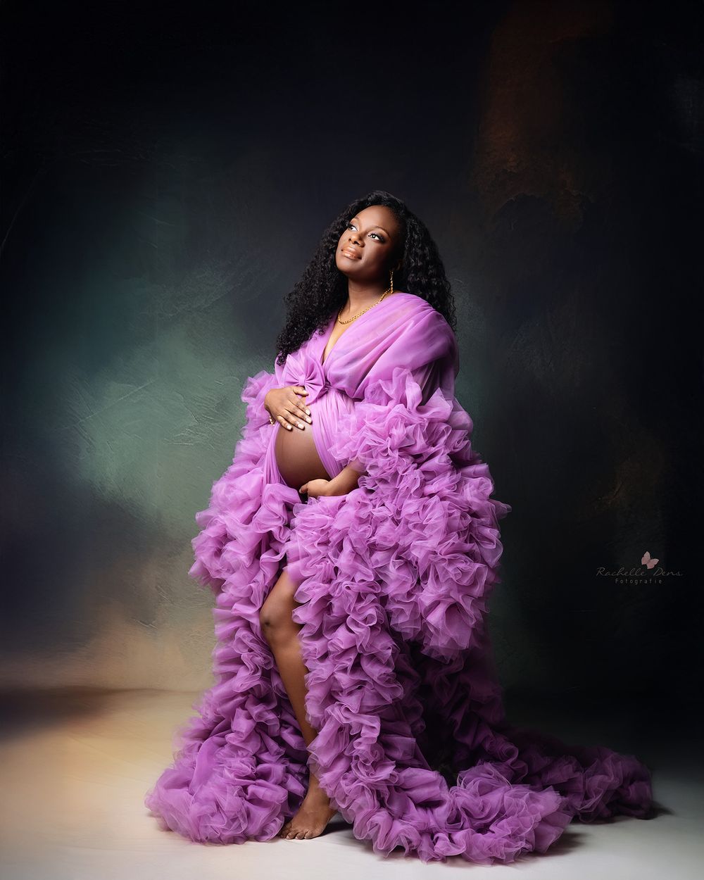 A maternity photoshoot features a flowing purple tulle gown against a dark dramatic backdrop with moody lighting.