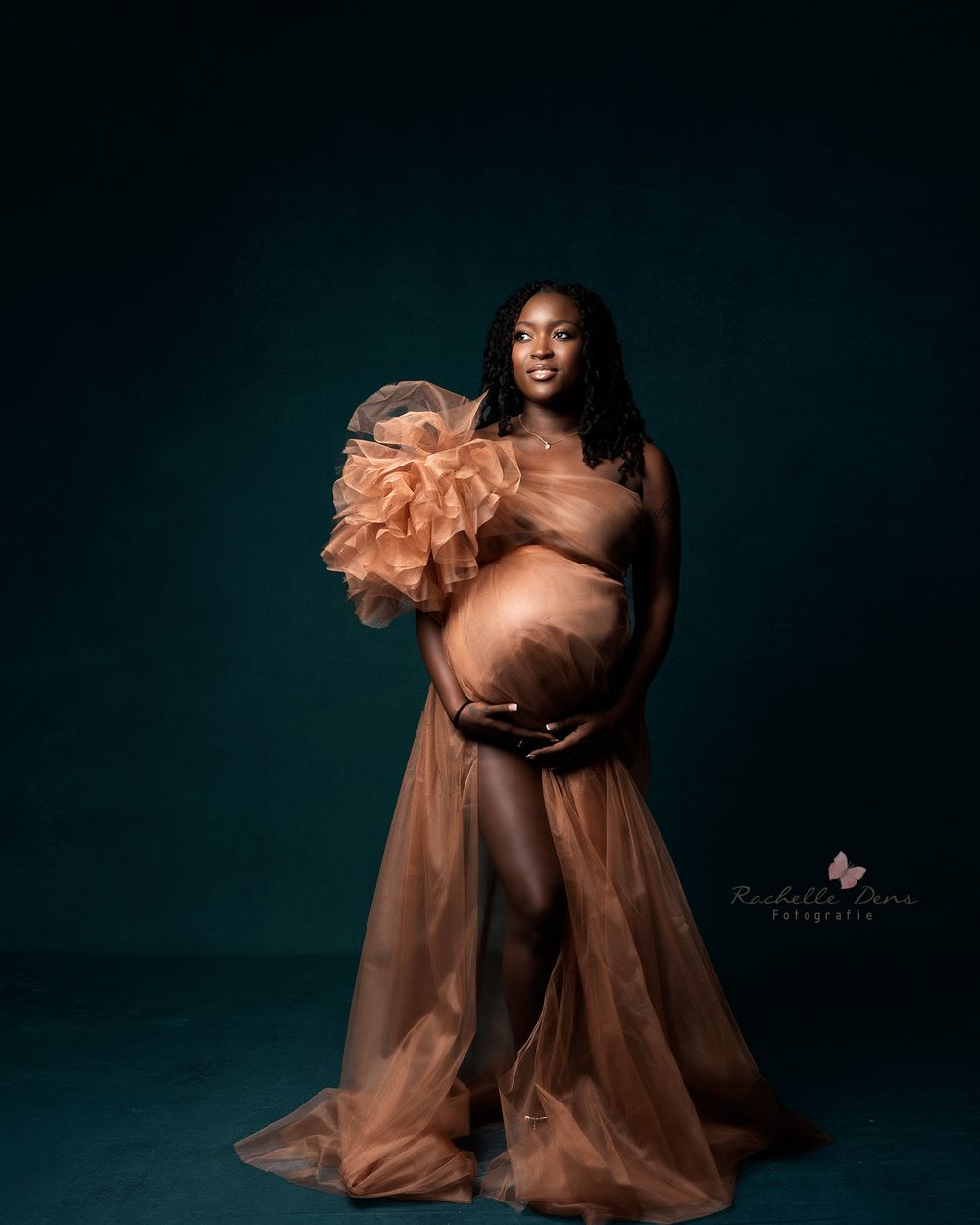 An elegant maternity photo showcasing a woman in a flowing peach-colored gown against a dark turquoise backdrop.